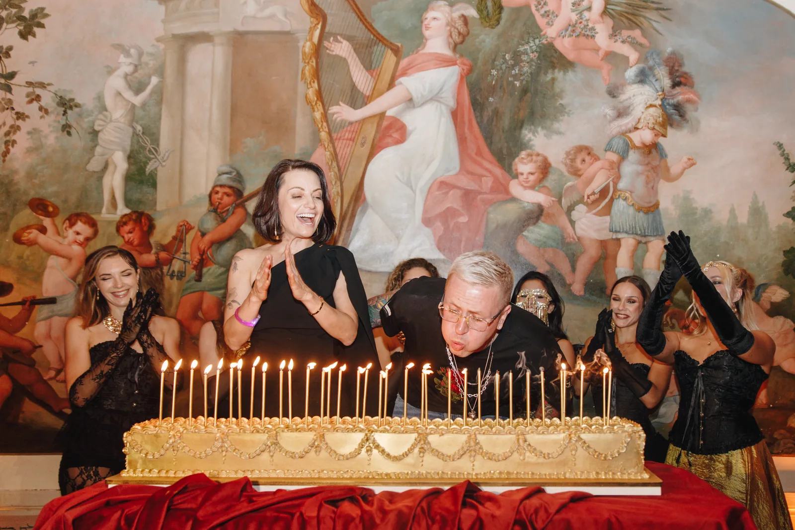 Masquerade milestone birthday at Palácio do Grilo in Lisbon — the guest of honour blowing out the candles on a gold-tiered cake against the palace's frescoed hall, produced by BTQ Events