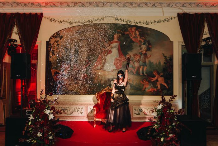 Masked performer on the red-carpet stage against the frescoed hall of Palácio do Grilo